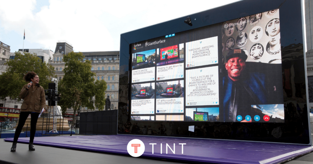 Woman with microphone standing in front of giant microsoft surface laptop, TINTmix from TINT social displays is on screen.