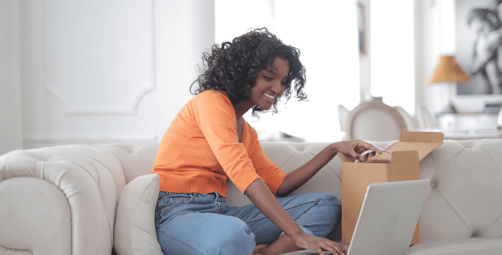 Woman sitting on sofa opening a box with a sample while typing on a laptop computer