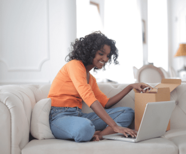 Woman sitting on sofa opening a box with a sample while typing on a laptop computer