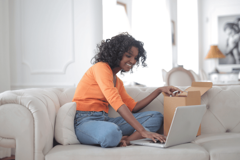 Woman sitting on sofa opening a box with a sample while typing on a laptop computer