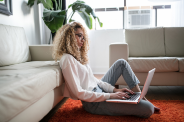 A woman sitting on the floor, leaned against her sofa with her laptop on her lap.