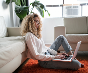 A woman sitting on the floor, leaned against her sofa with her laptop on her lap.