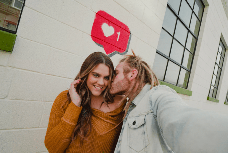 A man kissing a woman on the cheek with a like symbol behind them