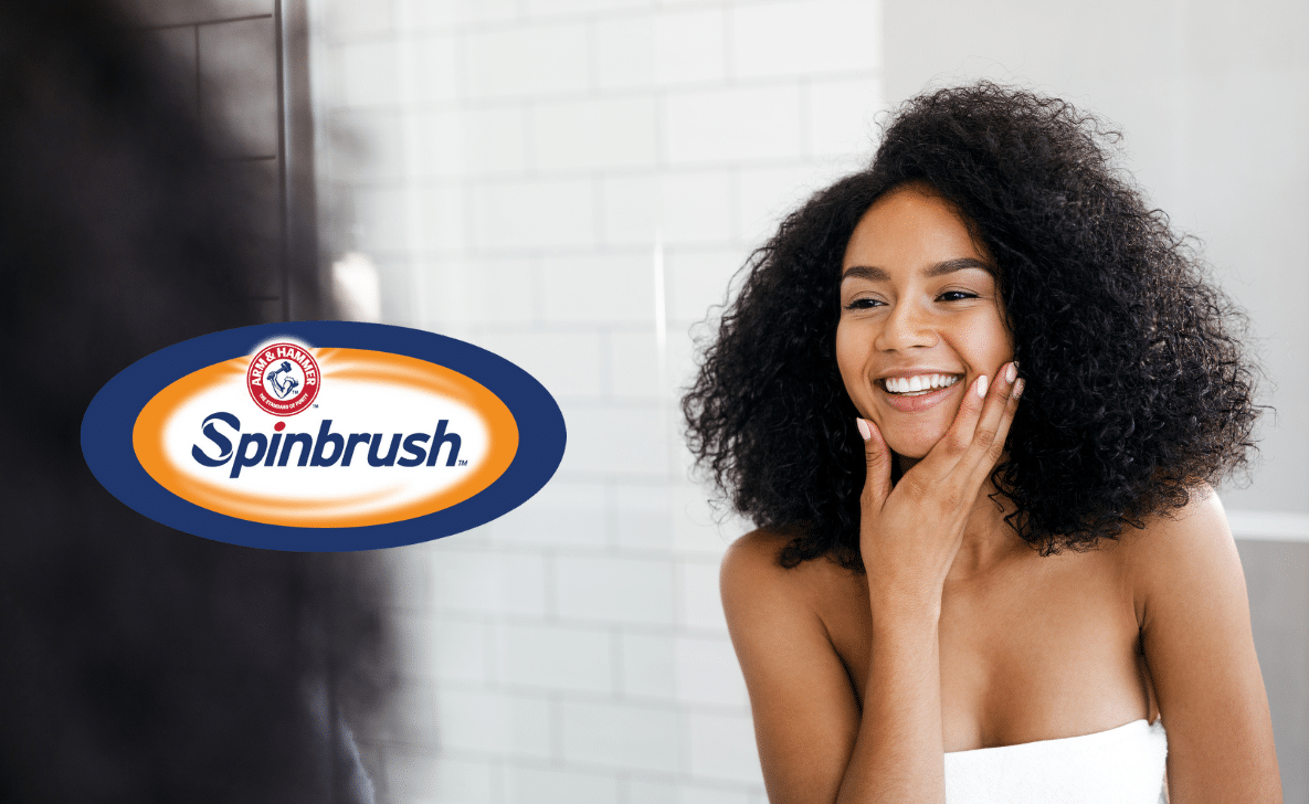 A woman smiling at her reflection in the mirror. ARM & HAMMER Spinbrush logo overlayed.